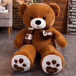 Brown teddy bear plush with a scarf, heart patch, and paw prints, sitting on a gray floor in a cozy room setting.