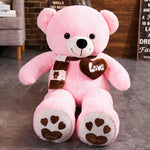 Pink teddy bear plush with a scarf, heart-shaped patch, and paw prints, sitting on a gray floor.