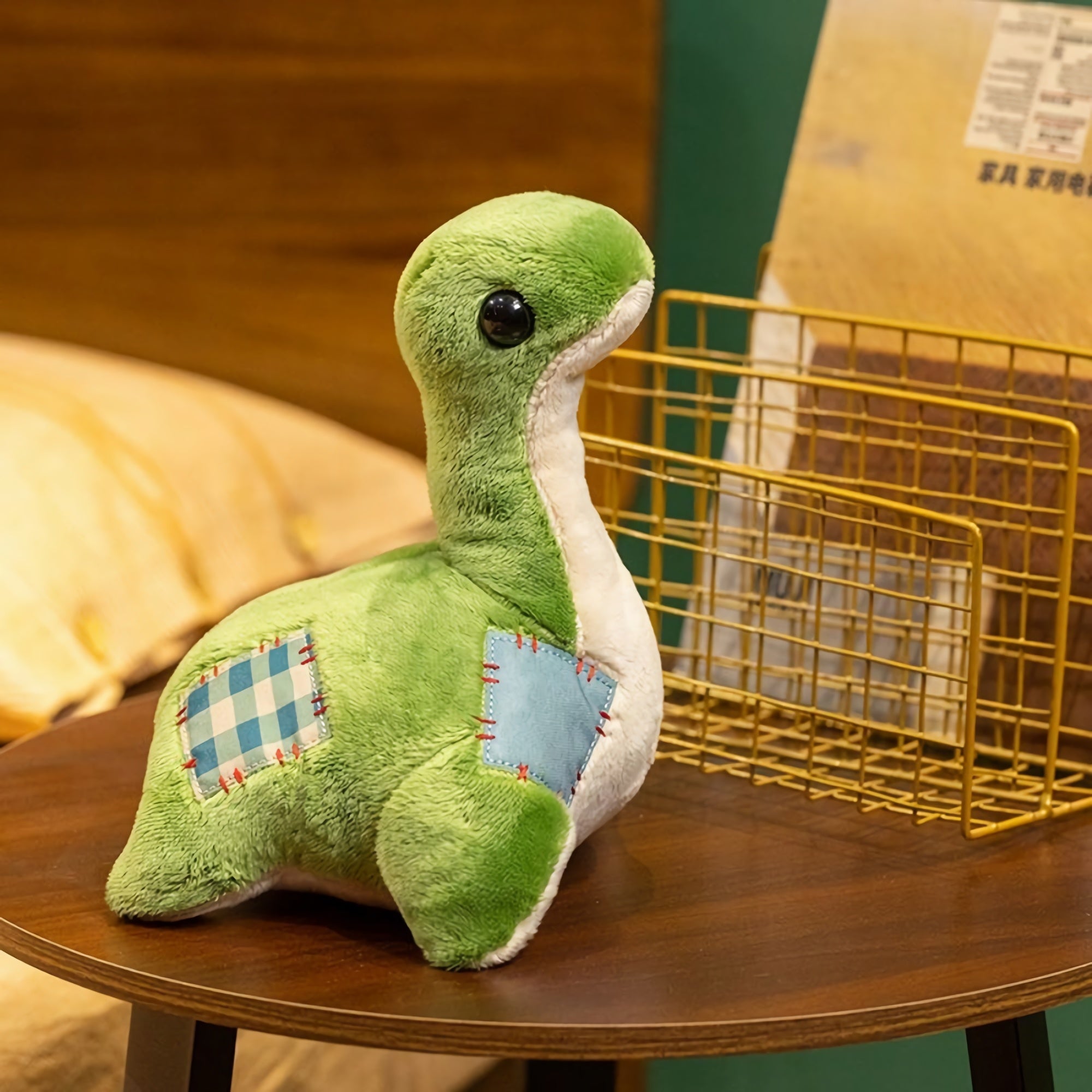 Green plush dinosaur with large eyes and patchwork details, sitting on a wooden table next to a wire basket.