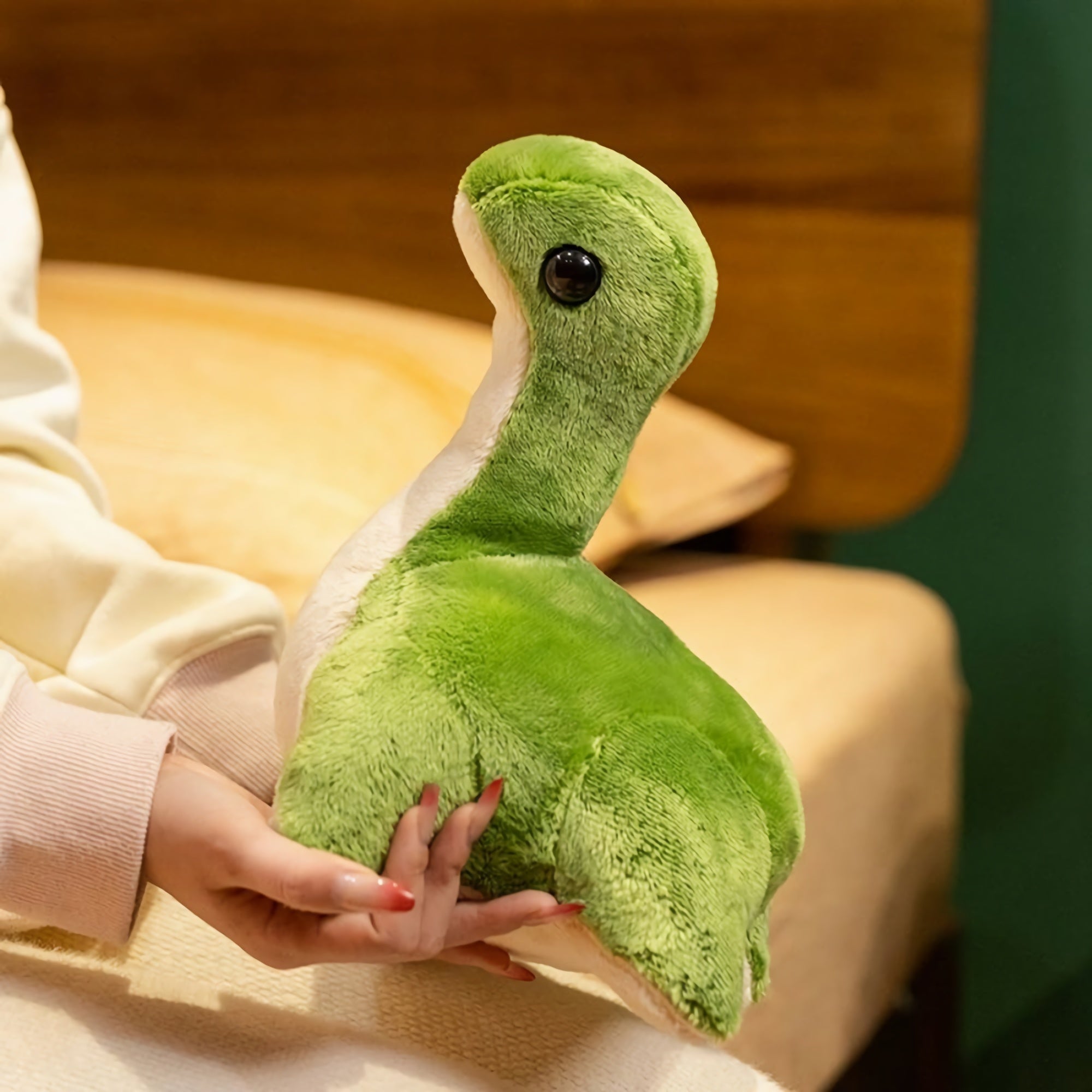 A person holds a green plush dinosaur with a white belly, sitting on a bed with a wooden headboard in the background.