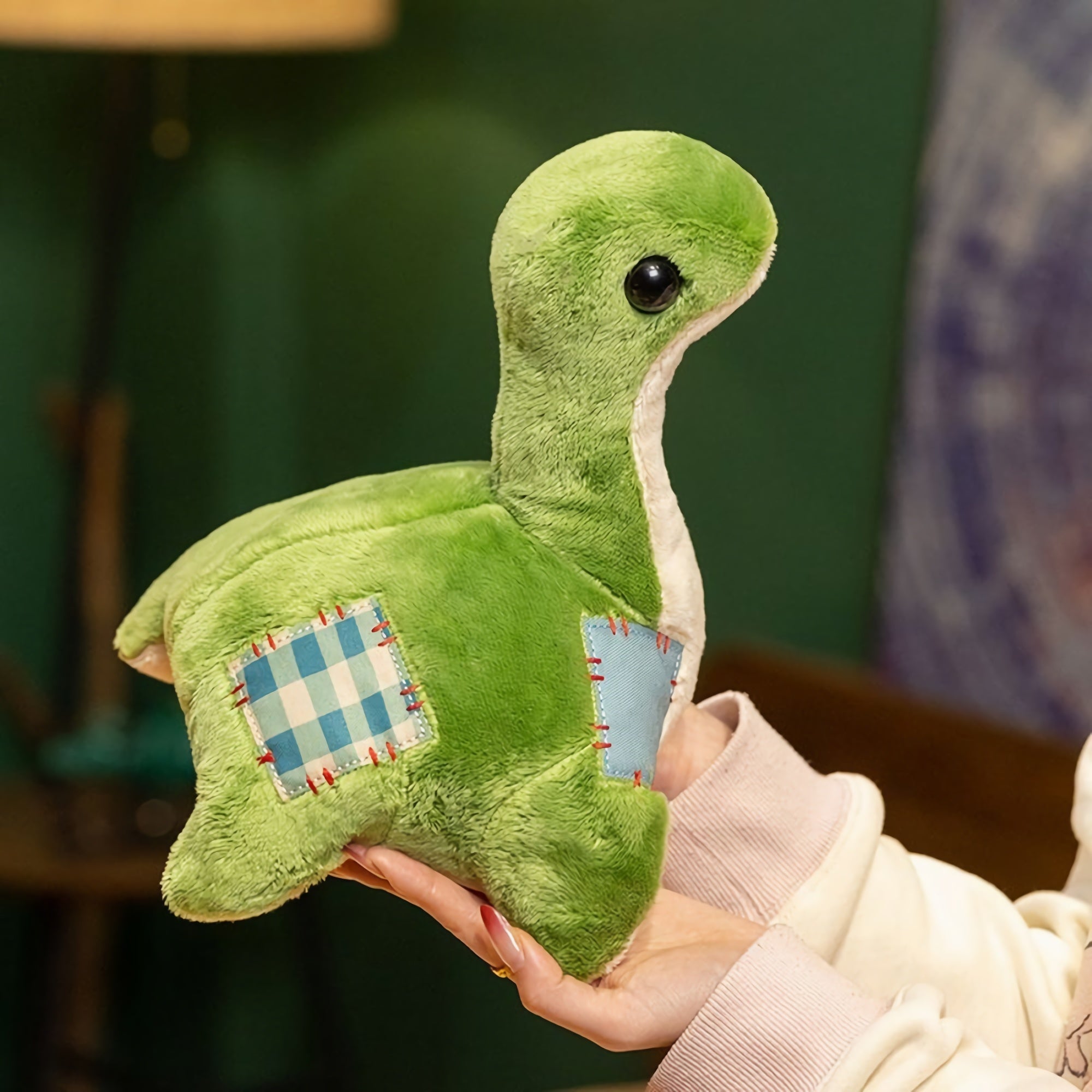 A green plush dinosaur with a round head and stitched patches, held in a person's hand against a dark background.