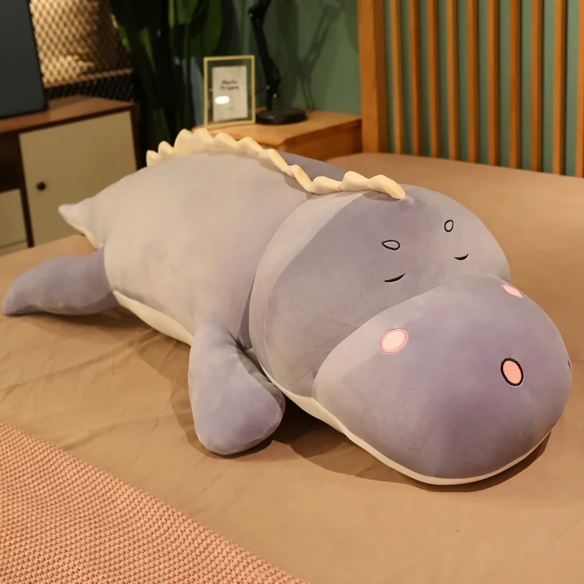 Plush dinosaur pillow in shades of purple, with a rounded body and cute facial features, resting on a bed.