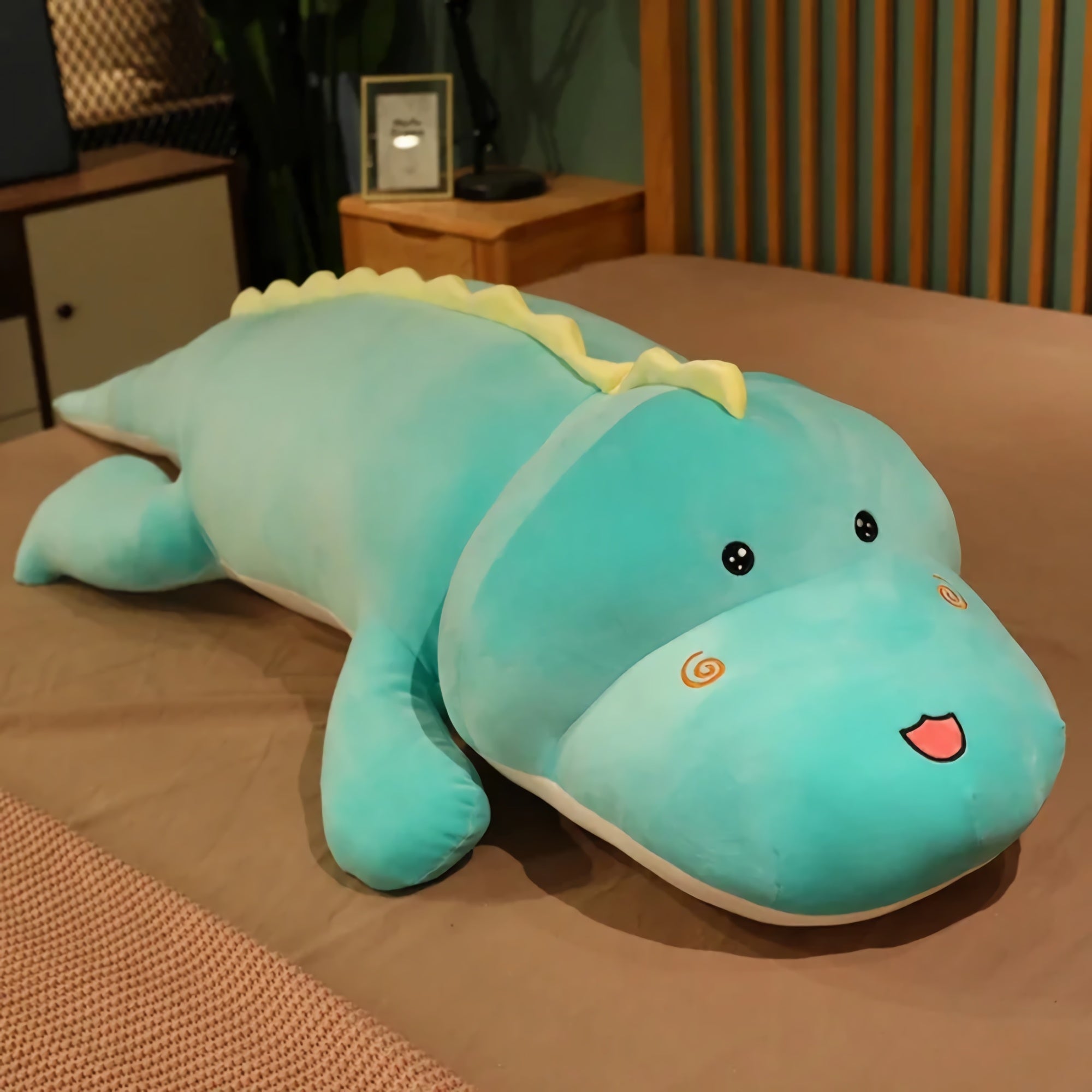 A large, soft turquoise dinosaur plush pillow with a smiling face and yellow spikes on its back.