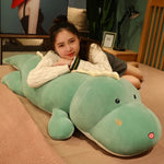 A girl rests her head on a large, soft green dinosaur plush pillow while lying on a bed.