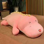 A pink dinosaur plush pillow with a smiling face and yellow spikes, resting on a brown surface.