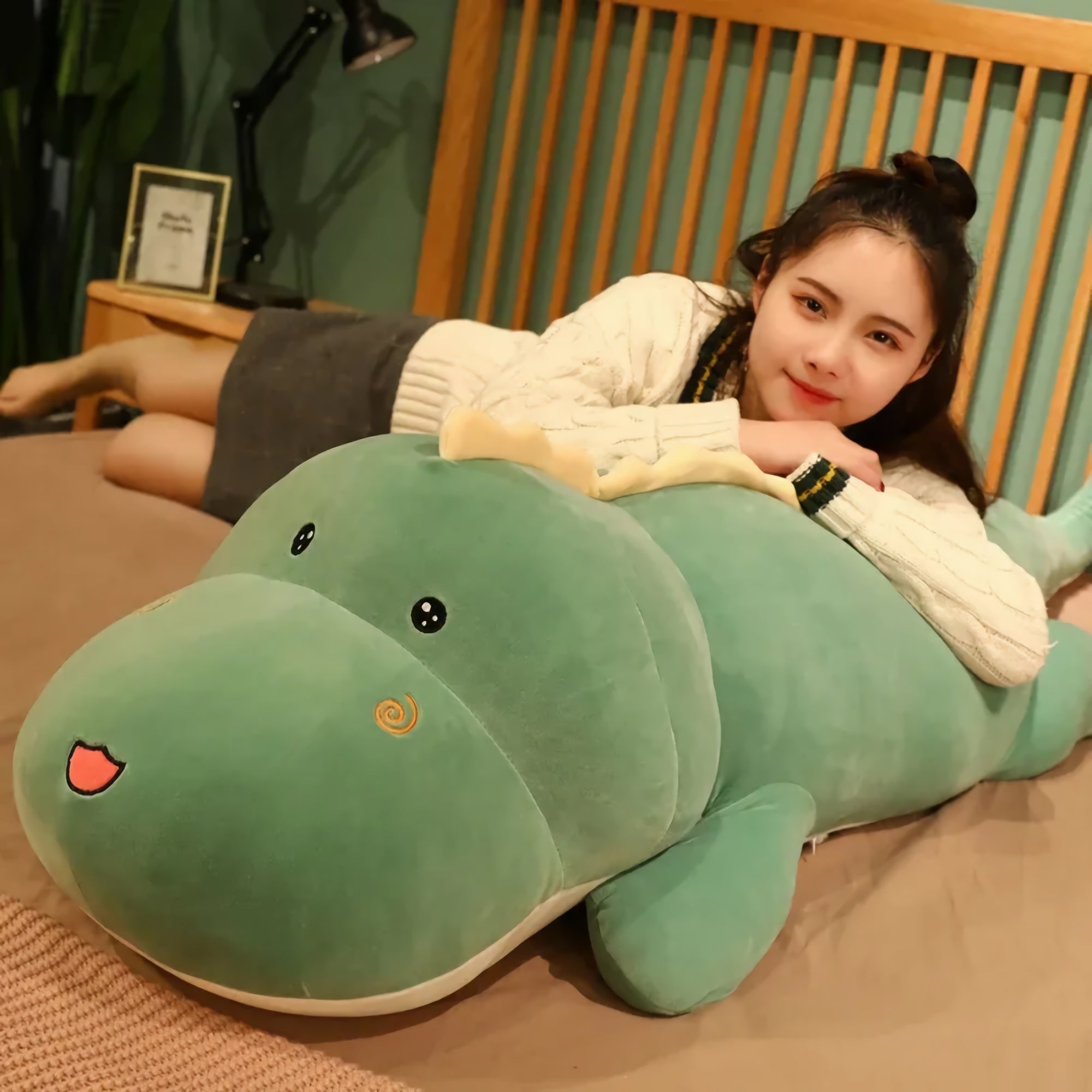 A person lies on a bed with a large green dinosaur plush pillow, smiling and resting on their arms.