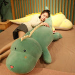 A person lies on a bed, hugging a large green dinosaur plush pillow, with a yellow dinosaur plush nearby.