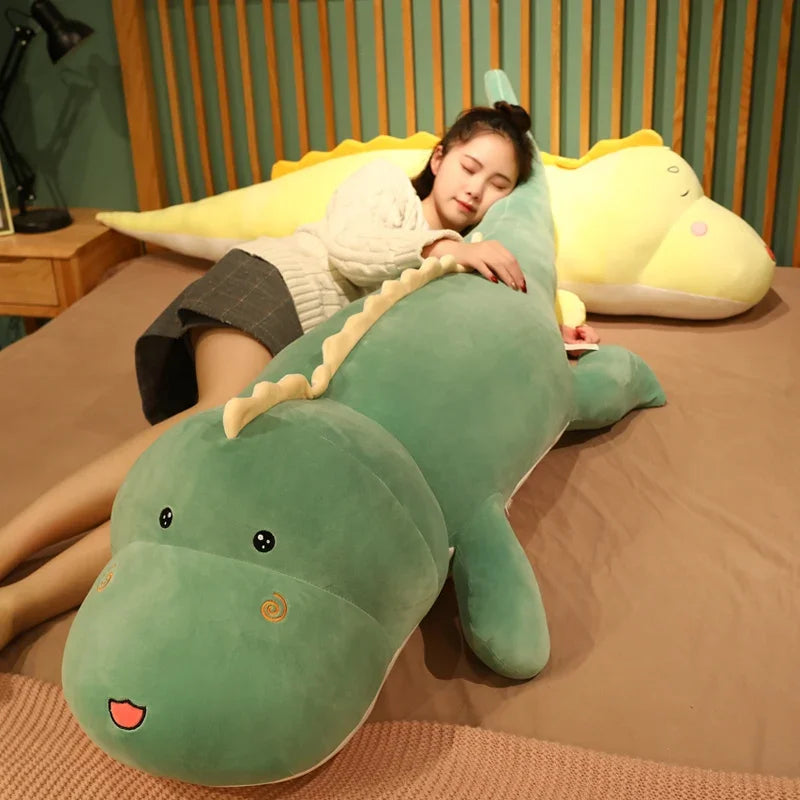 A person lies on a bed, hugging a large green dinosaur plush pillow, with a yellow dinosaur plush nearby.