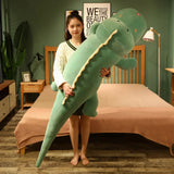A person holds a large green dinosaur plush pillow in a bedroom setting.
