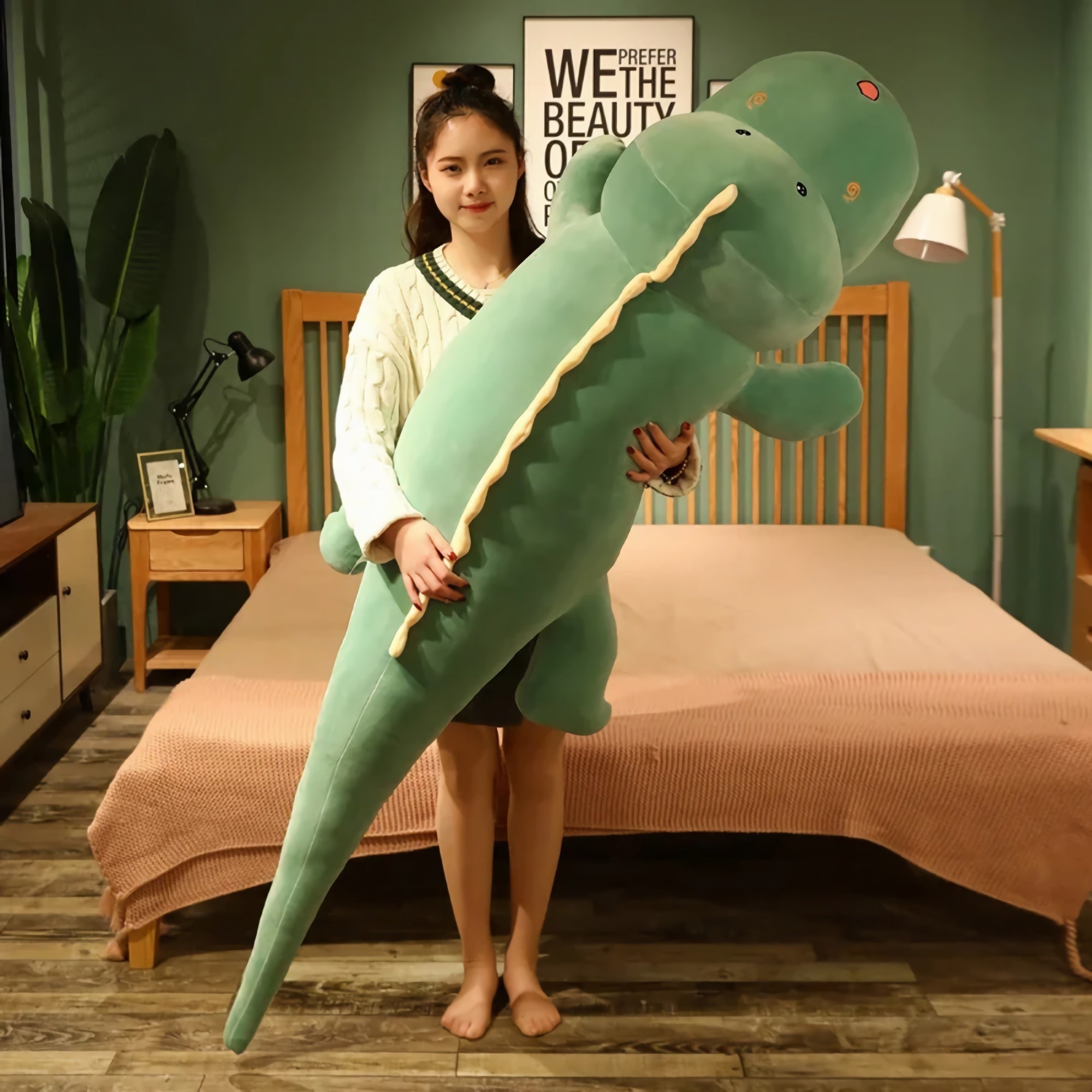 A person holds a large green dinosaur plush pillow in a bedroom setting.