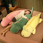 A person sleeps on a bed surrounded by three large, colorful dinosaur plush pillows in pink, green, and yellow.