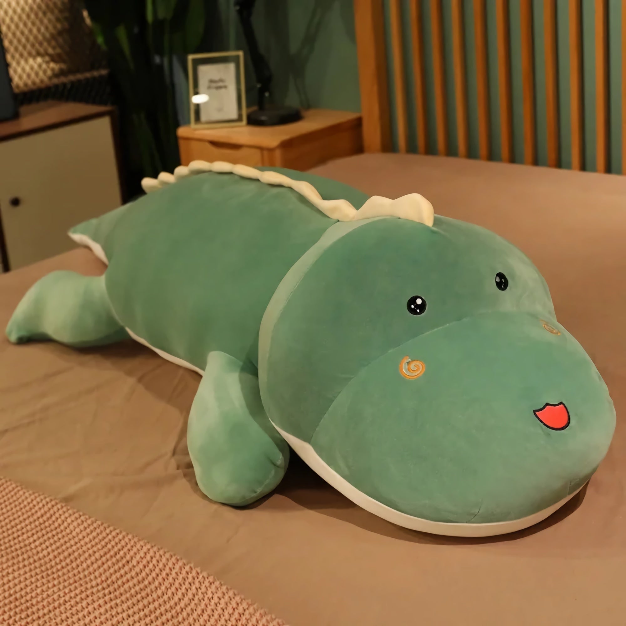Kawaii long dinosaur plush pillow in green with a smiling face, placed on a bed.
