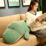 A person sits on a couch, reading a book next to a large green dinosaur plush pillow.