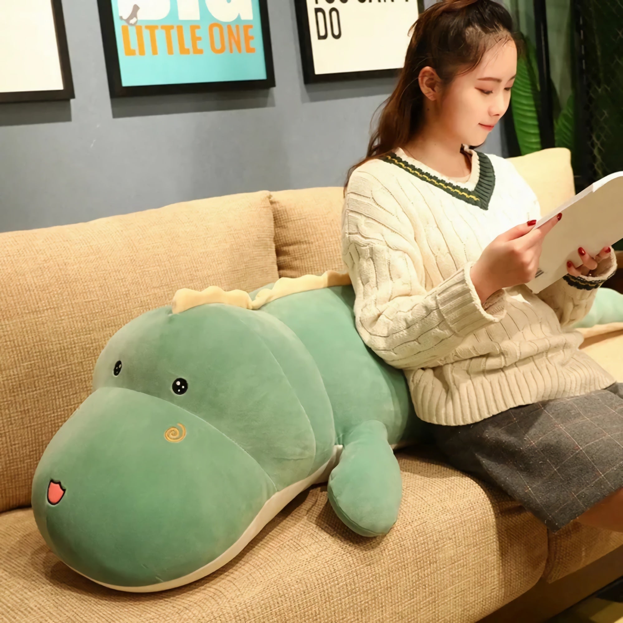 A person sits on a couch, reading a book next to a large green dinosaur plush pillow.