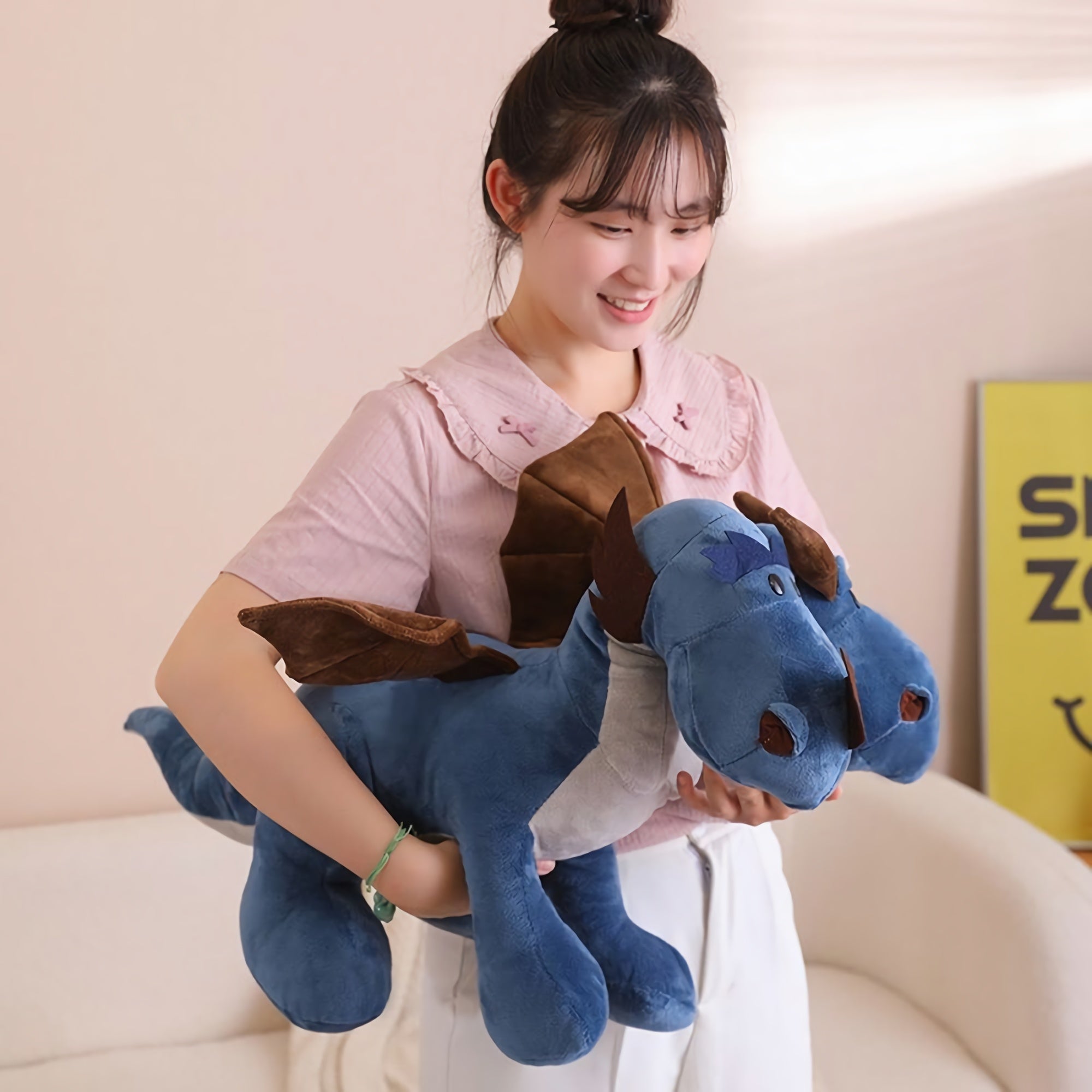 A person holds a large blue plush dragon with brown wings, smiling in a bright room.