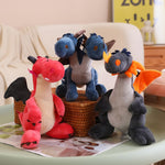 Three plush dragon toys in red, blue, and gray with wings, sitting on a table with magazines.