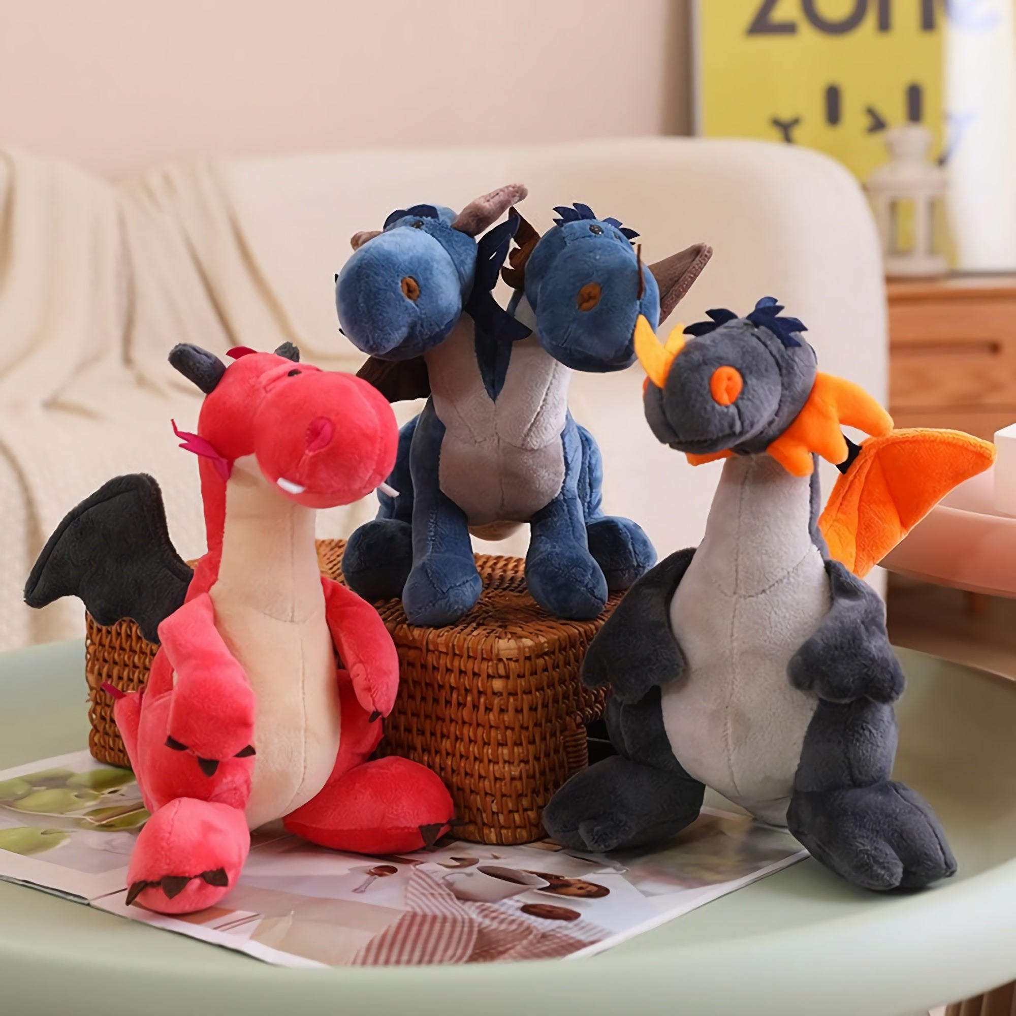 Three plush dragon toys in red, blue, and gray with wings, sitting on a table with magazines.