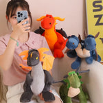 A person holds a plush dragon while surrounded by colorful plush dragons and dinosaurs on a couch.