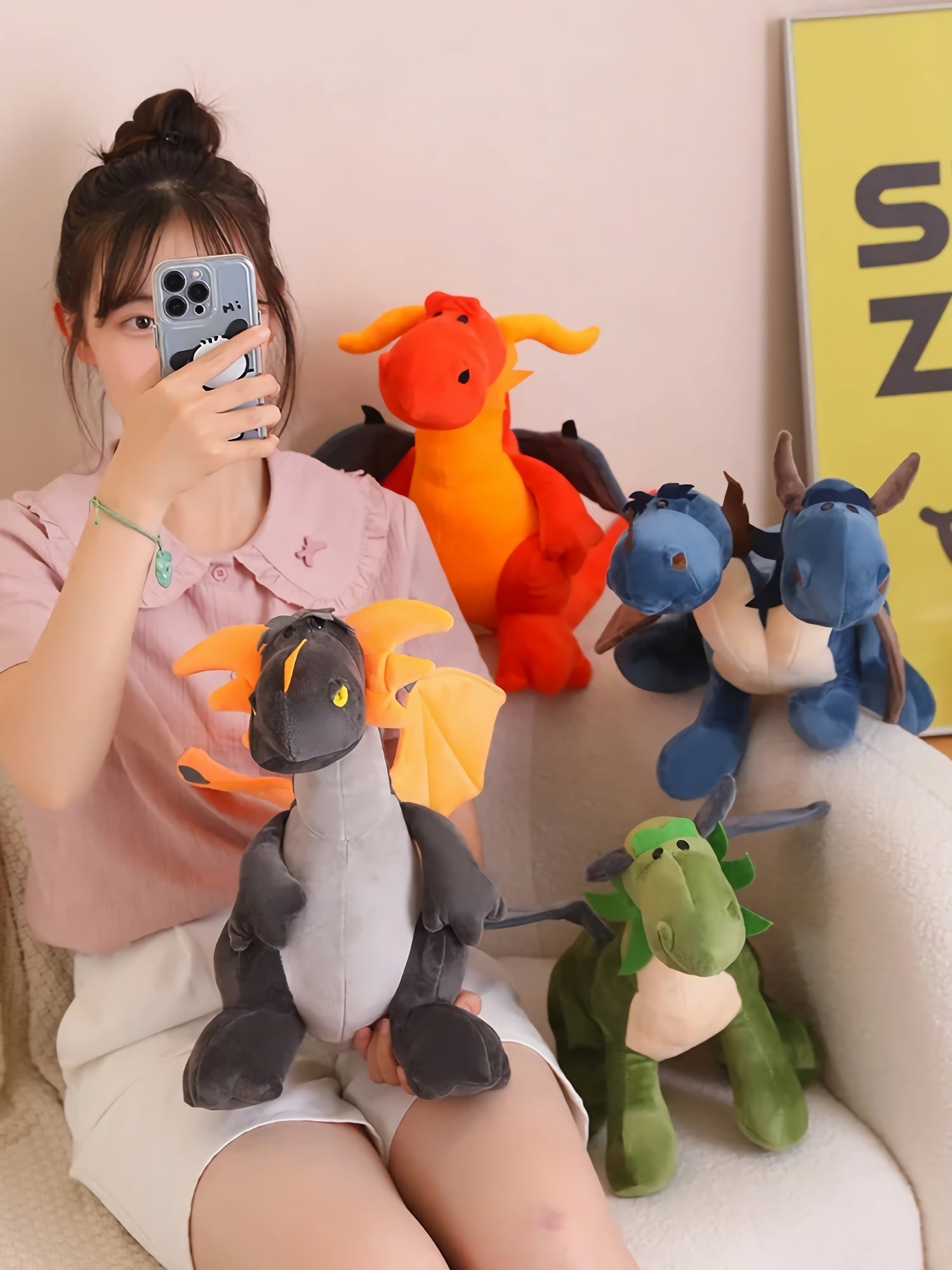 A person holds a plush dragon while surrounded by colorful plush dragons and dinosaurs on a couch.