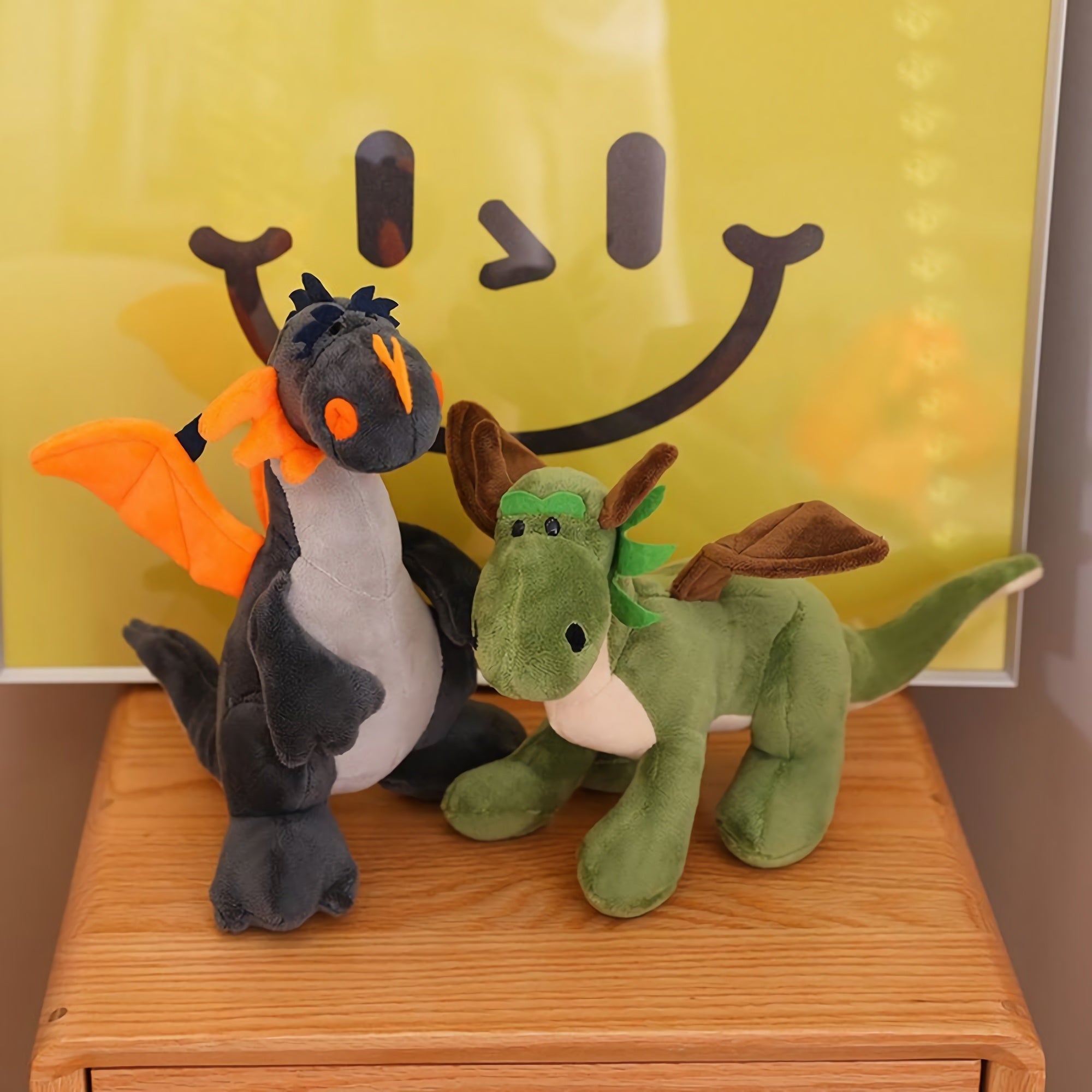 Two plush toys, a black and orange dragon and a green dinosaur with wings, on a wooden surface in front of a yellow poster.