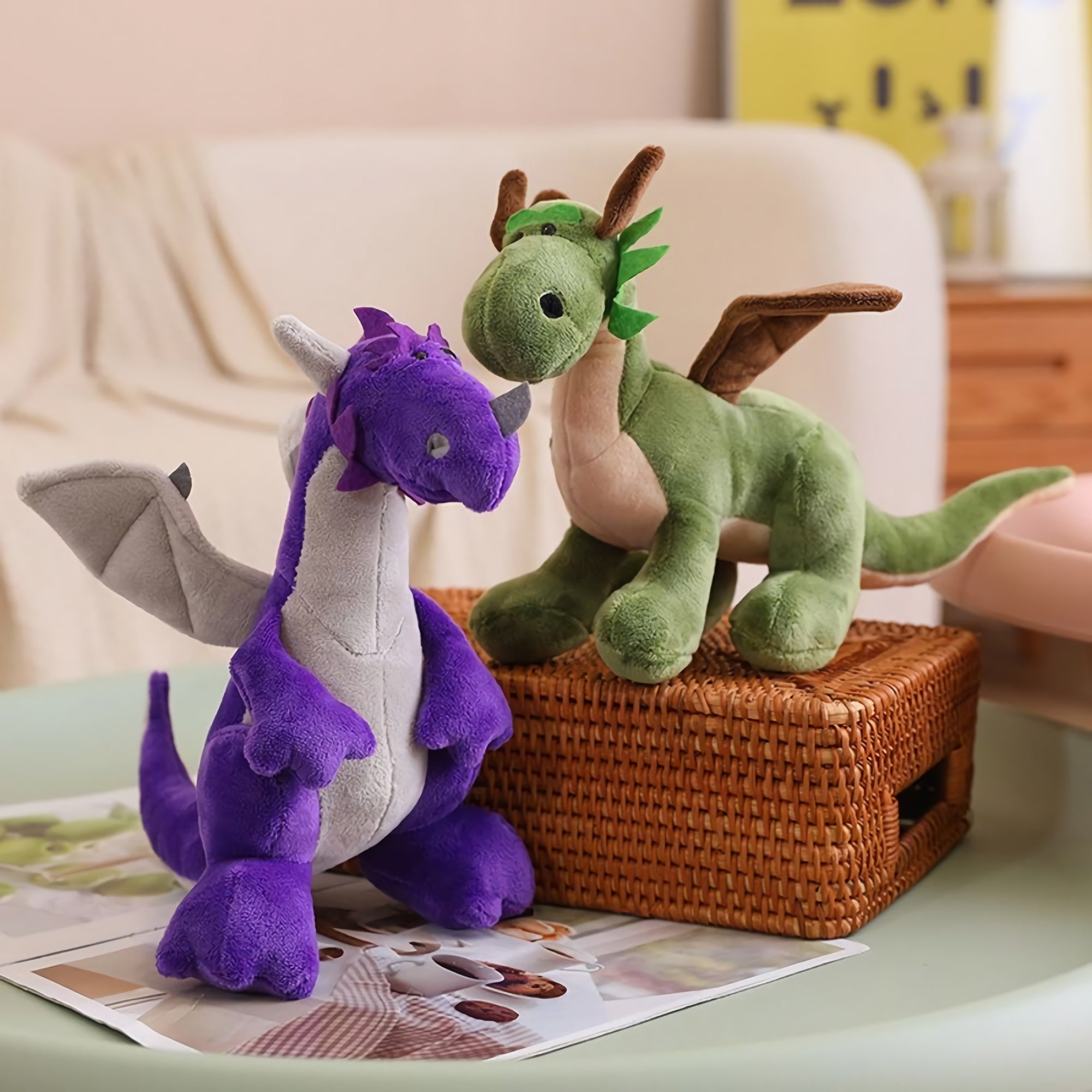 Two plush toys: a purple dragon with gray wings and a green dinosaur with brown wings, sitting on a table.