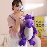 A person holds a purple plush dragon with gray accents, smiling while taking a selfie.