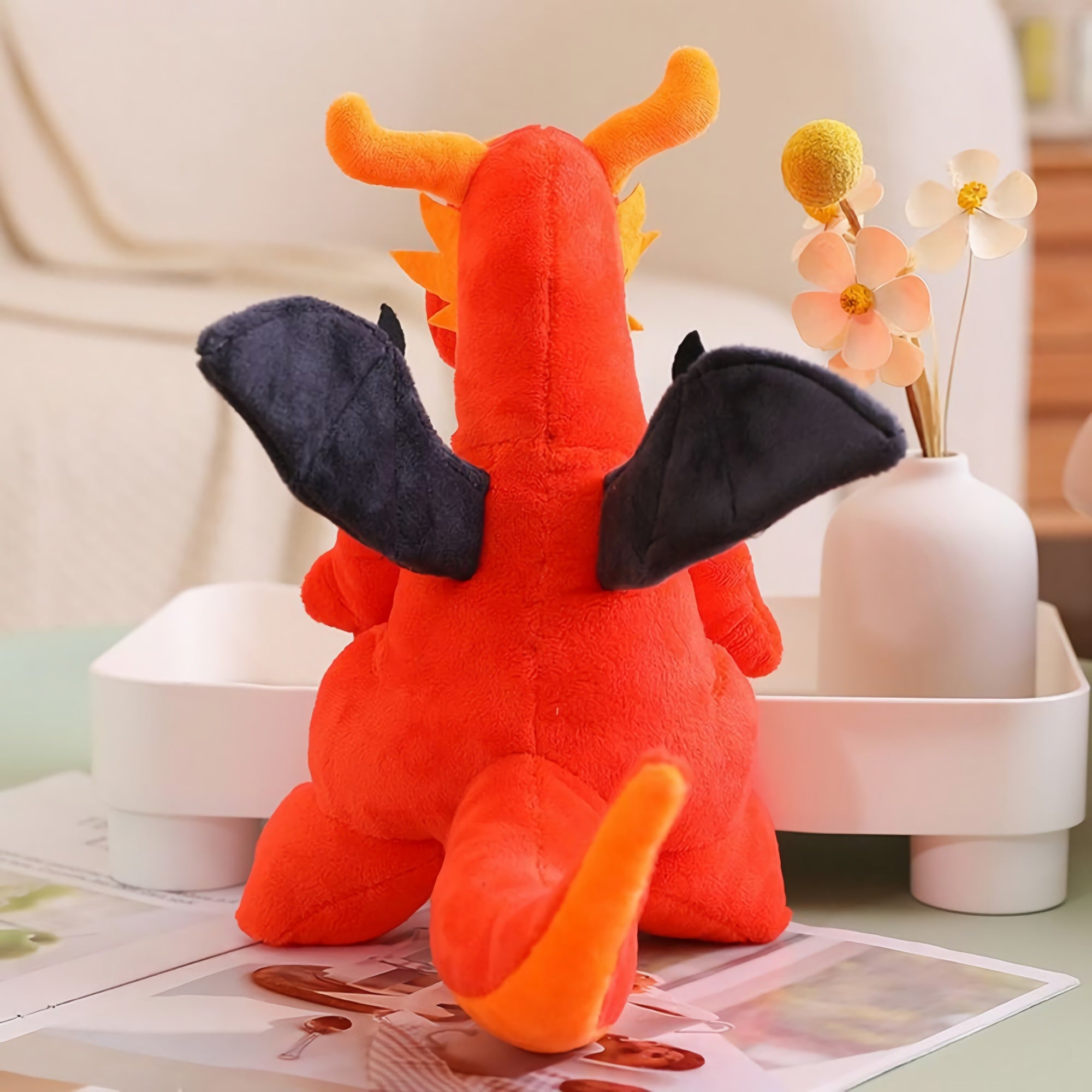 Plush toy of a winged dragon in red with black wings, sitting on a table beside a vase and magazines.