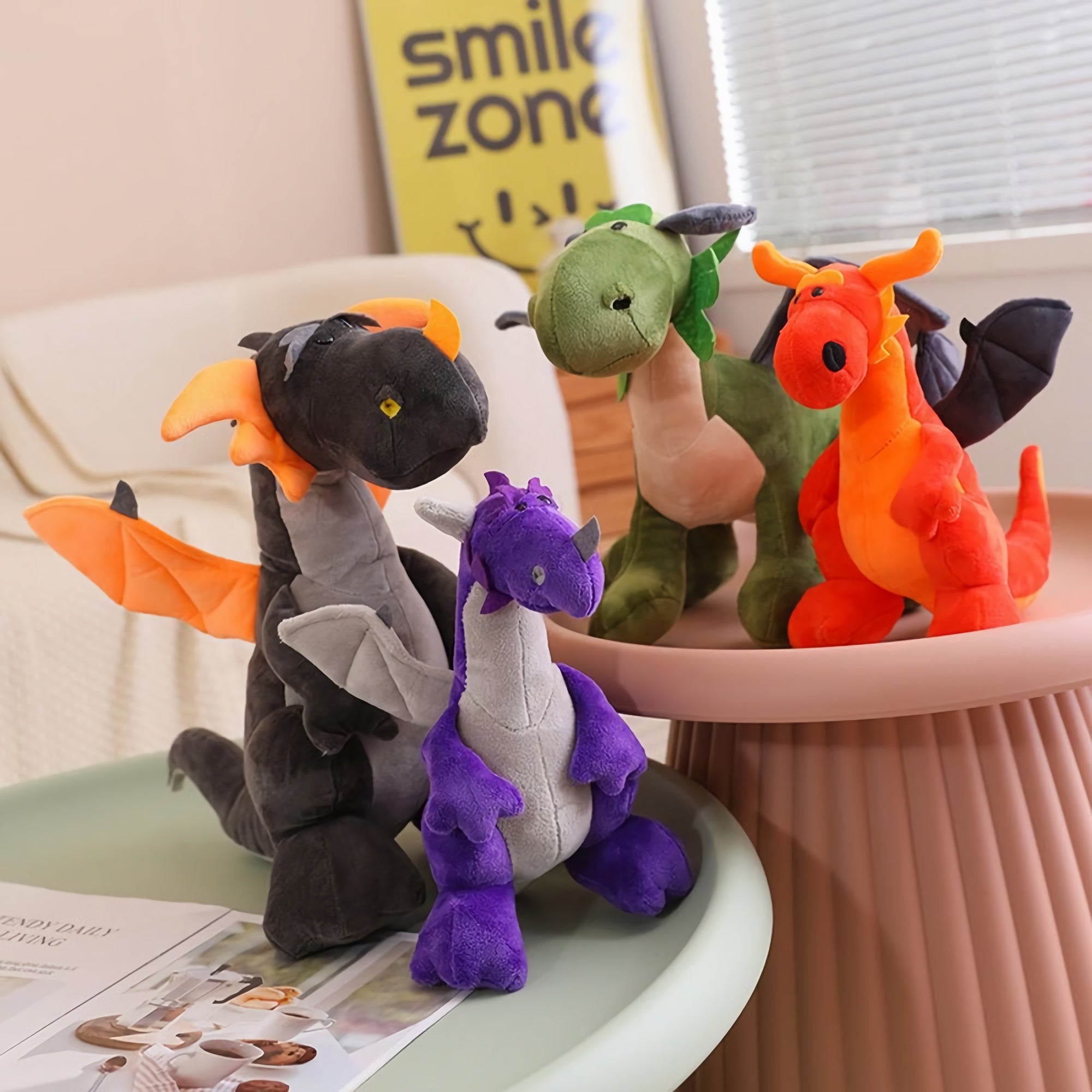 Four plush dragon and dinosaur toys in various colors, sitting on a table with a magazine nearby.
