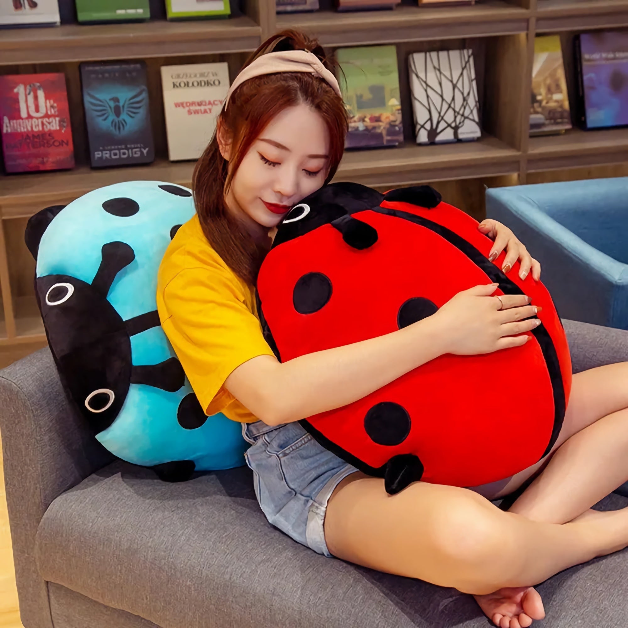 A person sits on a couch, hugging colorful ladybug plush pillows, one red and one blue, in a cozy setting.