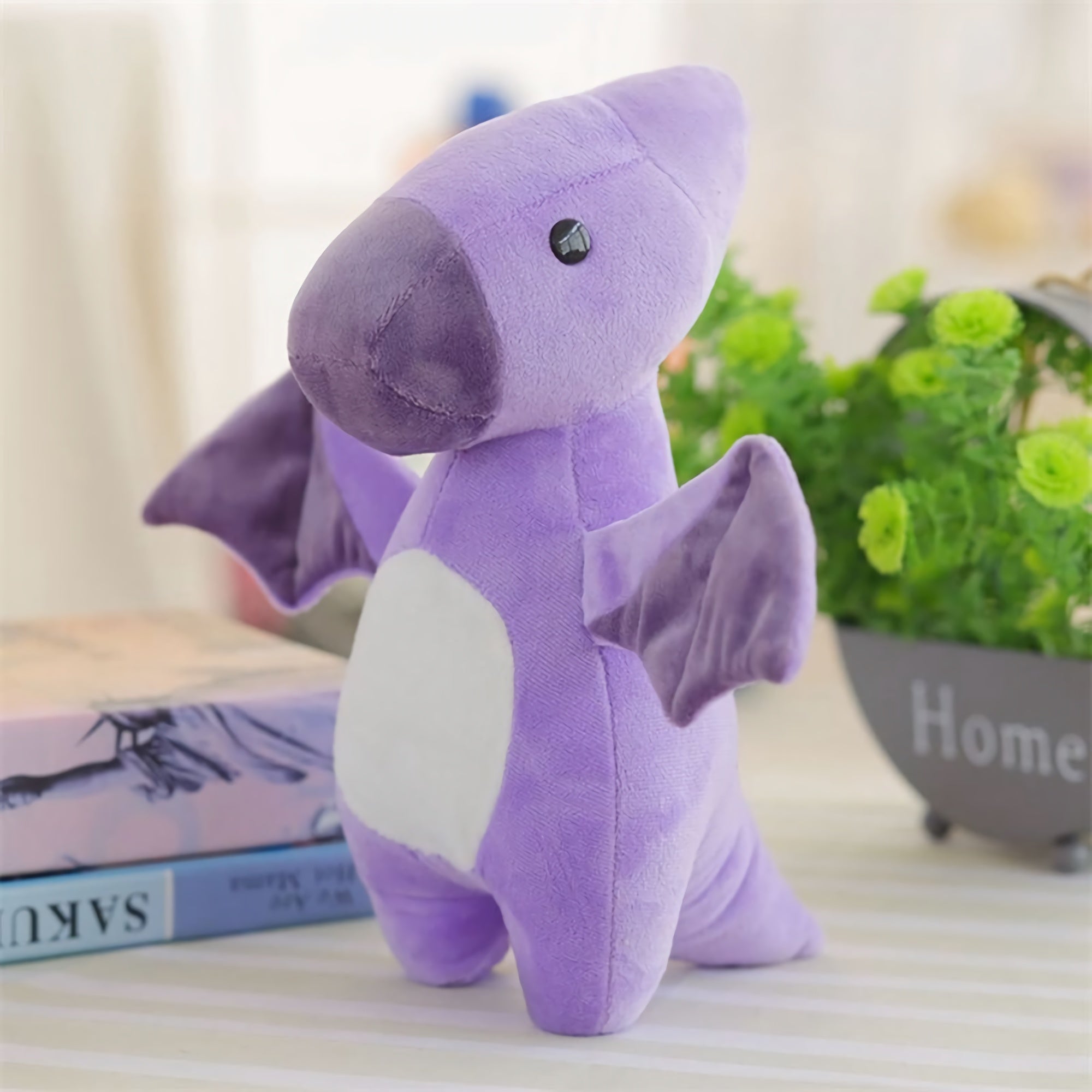 Pastel purple dinosaur plush toy with wings, sitting on a table near books and a small plant.
