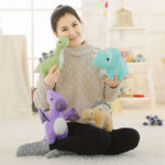 A person sitting on a rug holds four colorful dinosaur plush toys in a bright room.