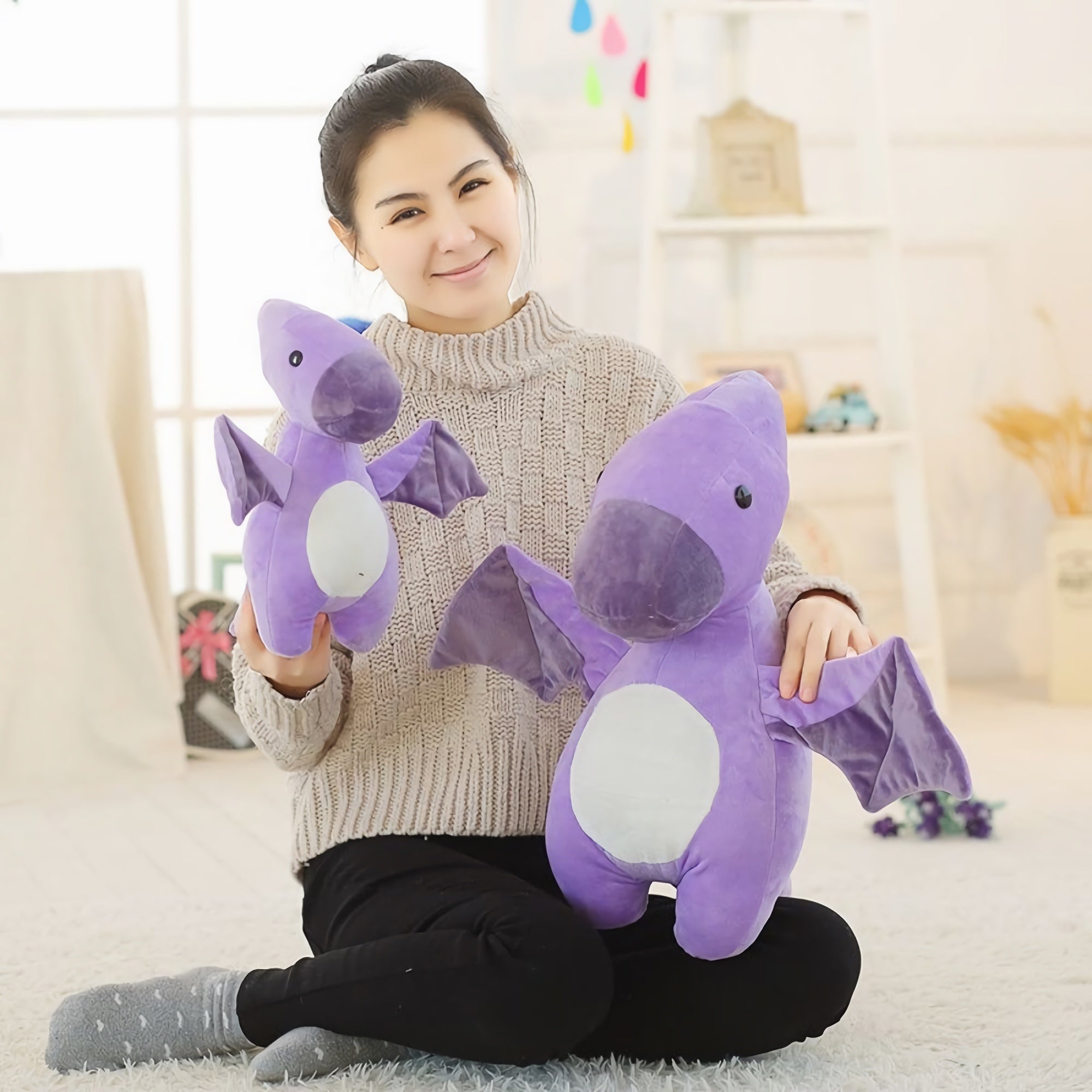 A woman sits on the floor holding two purple dinosaur plush toys, smiling in a bright, cozy room.