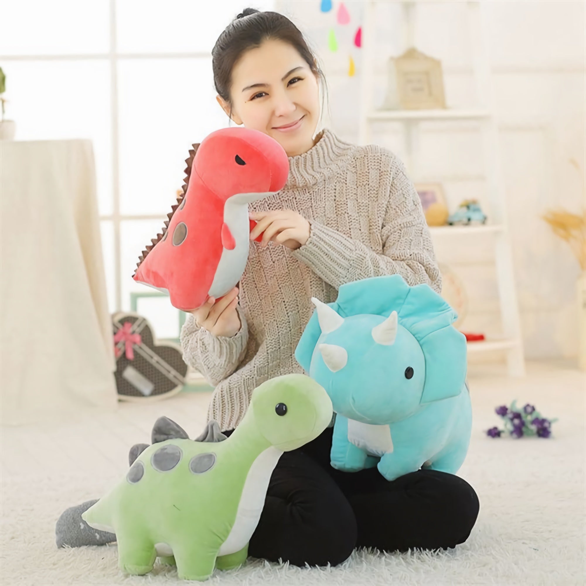 A woman holds a red dinosaur plush toy, with a green and blue dinosaur plush toy beside her on a soft floor.
