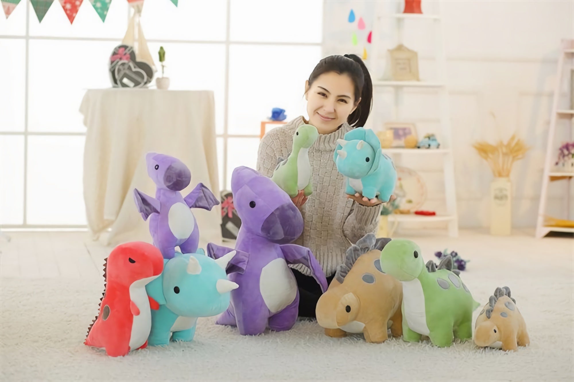 A person holds two pastel dinosaur plush toys surrounded by several others on a soft surface in a bright room.