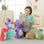 A person holds two pastel dinosaur plush toys surrounded by several others on a soft surface in a bright room.