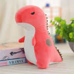 Pastel pink dinosaur plush toy with a white belly and small spikes, sitting beside books and a plant.