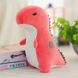 Pastel pink dinosaur plush toy with a white belly and small spikes, sitting beside books and a plant.