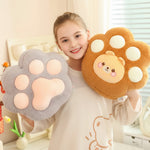 A girl holds two plush paw-shaped pillows, one gray and one brown, with a smiling bear face on the brown one.