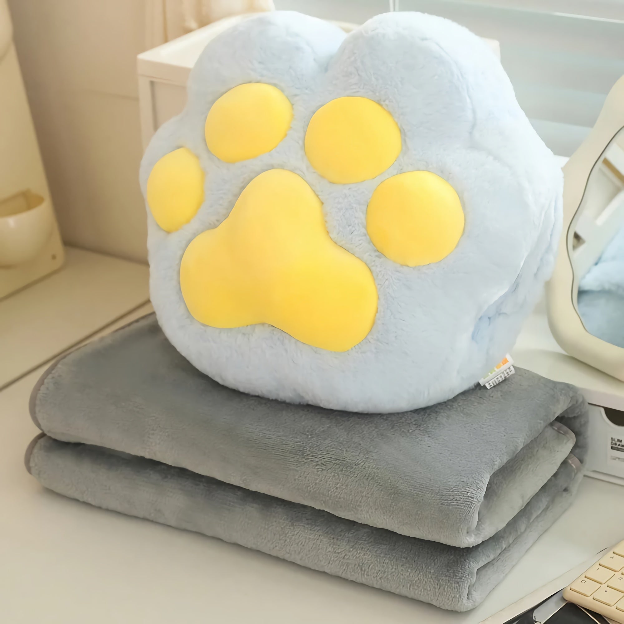 A blue plush pillow shaped like a paw sits on top of two folded gray blankets on a table.