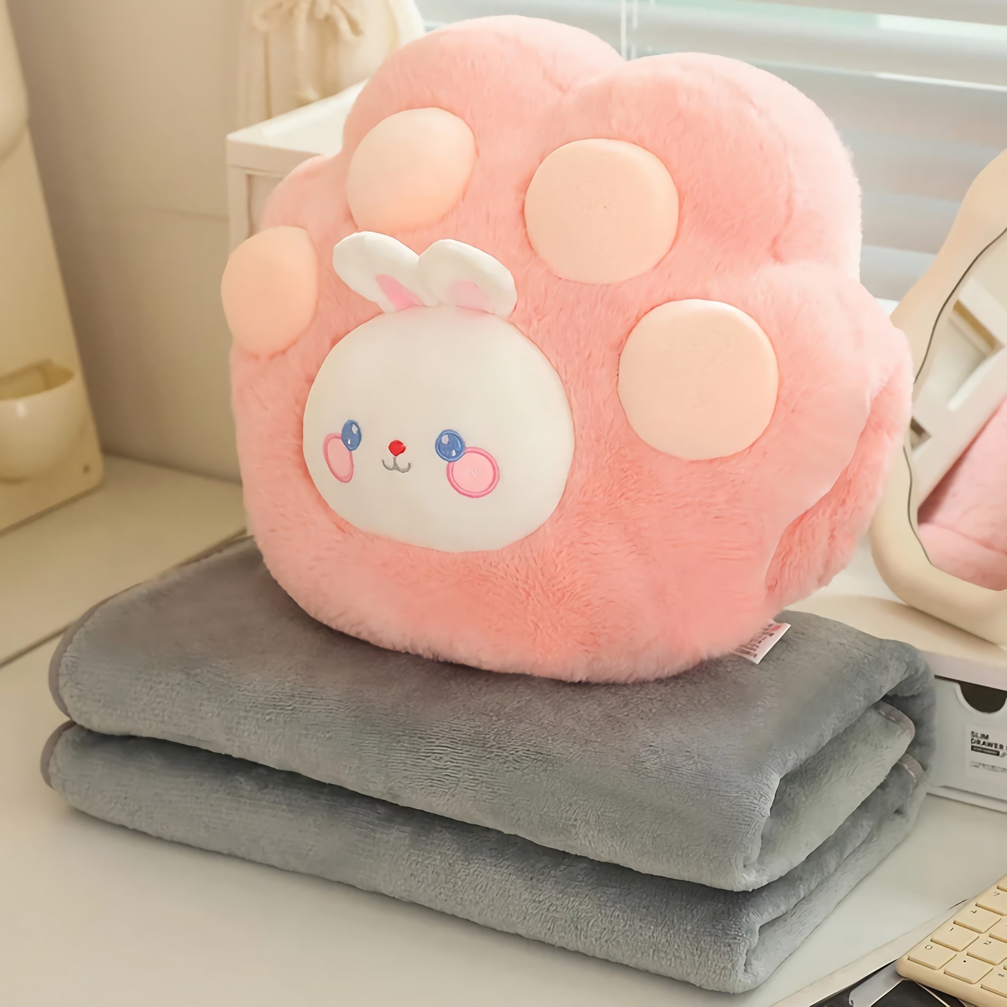 Pink plush paw-shaped pillow with a bunny face on top of two folded gray blankets.