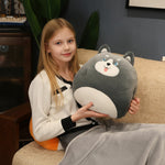 A girl sits on a couch holding a round plush pillow shaped like a gray animal, wearing a cozy outfit.