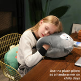 A child rests her head on a plush gray pillow while sitting at a table, with a green plush toy nearby.