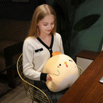 A girl sits in a chair holding a round, plush yellow chick pillow, smiling softly while looking down.