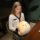 A girl sits in a chair holding a round, plush yellow chick pillow, smiling softly while looking down.