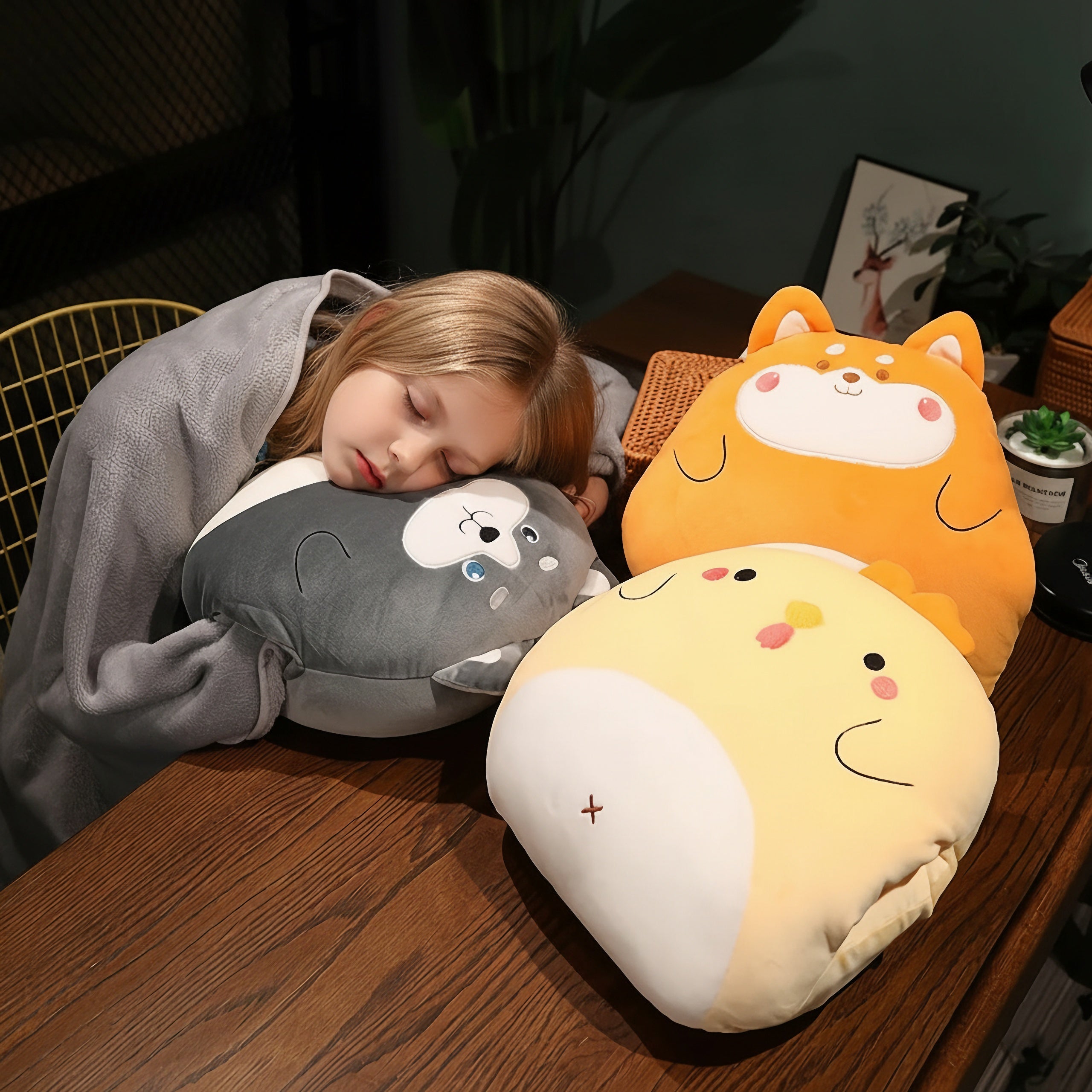 Child sleeping on a table with three plush pillows shaped like animals, wrapped in a gray blanket.