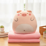 A plush pig-shaped pillow sits on a folded pink blanket on a wooden table.