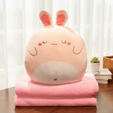 A round, pink plush bunny pillow sits atop two folded pink blankets on a wooden table.