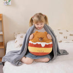 A child sits on a bed, wrapped in a gray blanket, holding a plush pillow shaped like a bear.