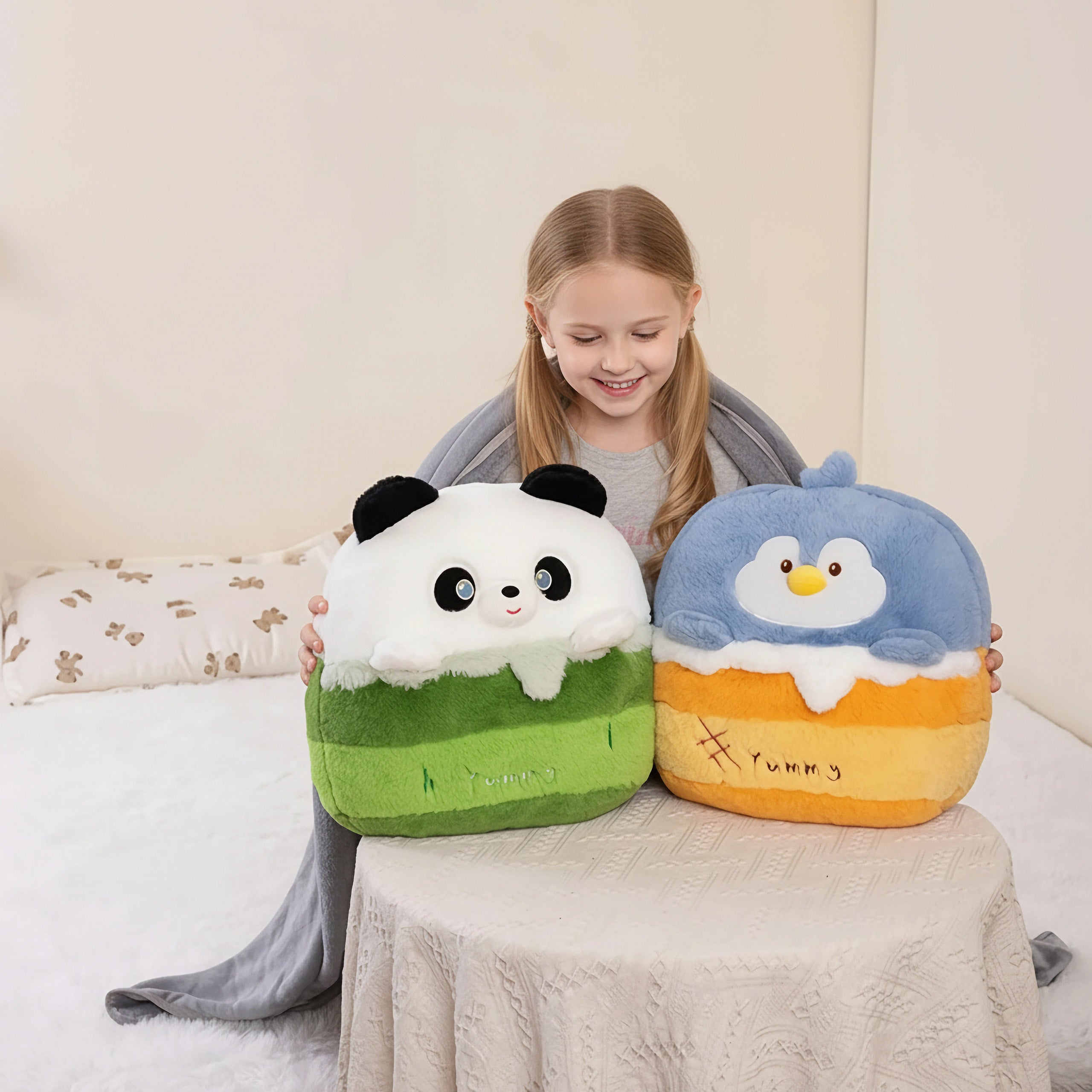 A girl smiles while holding two plush pillows shaped like a panda and a bird, sitting on a bed with a blanket.