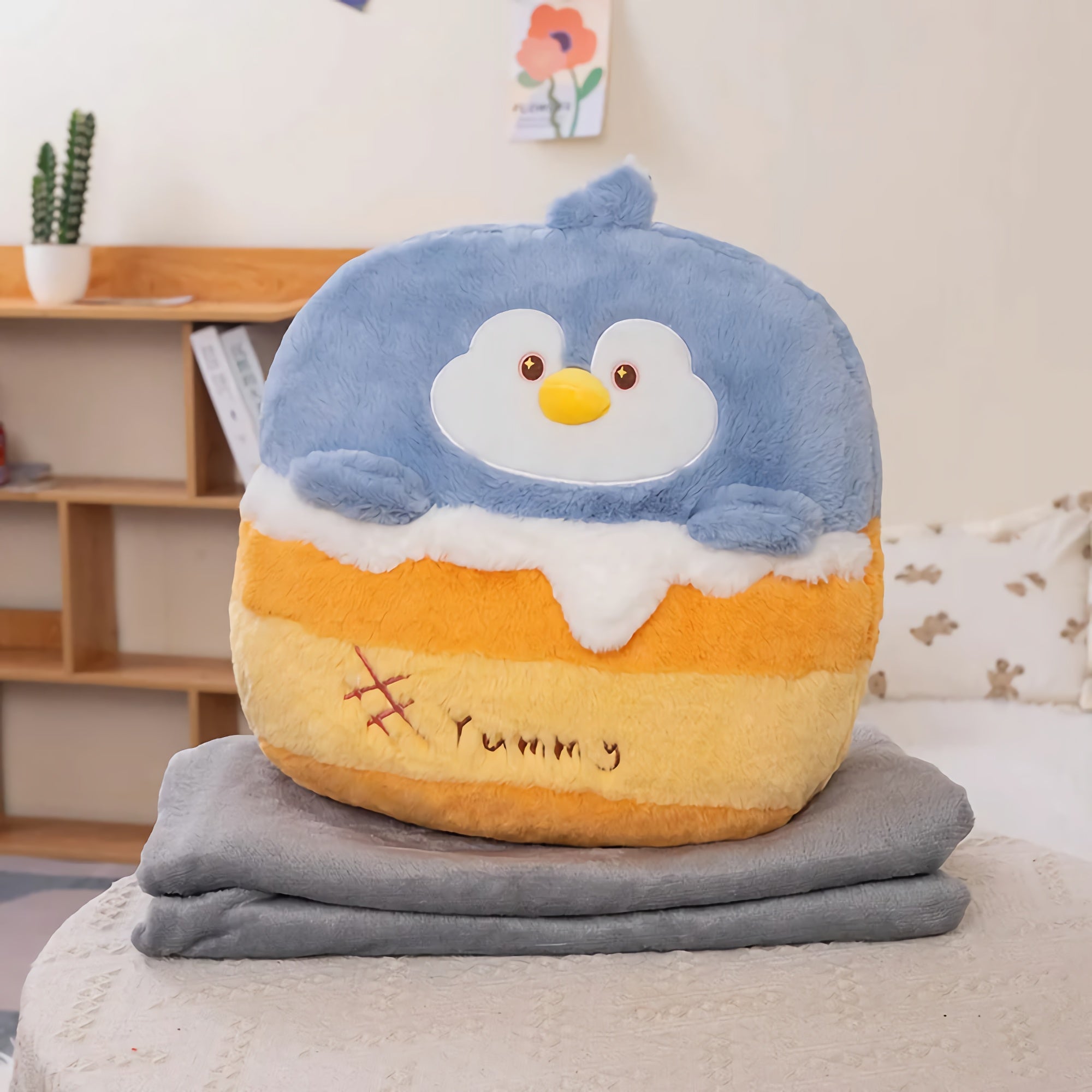 Plush penguin pillow with blue and orange colors on a folded gray blanket in a cozy room setting.